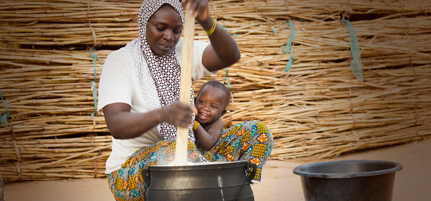 En bekymret mor sitter på huk og rører i ei stor gryte mens et lite barn henger smilende og forventningsfullt over låret hennes.